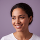 Portrait of a smiling woman in her late 20s, with a warm and joyful expression, against a dusty lilac background.